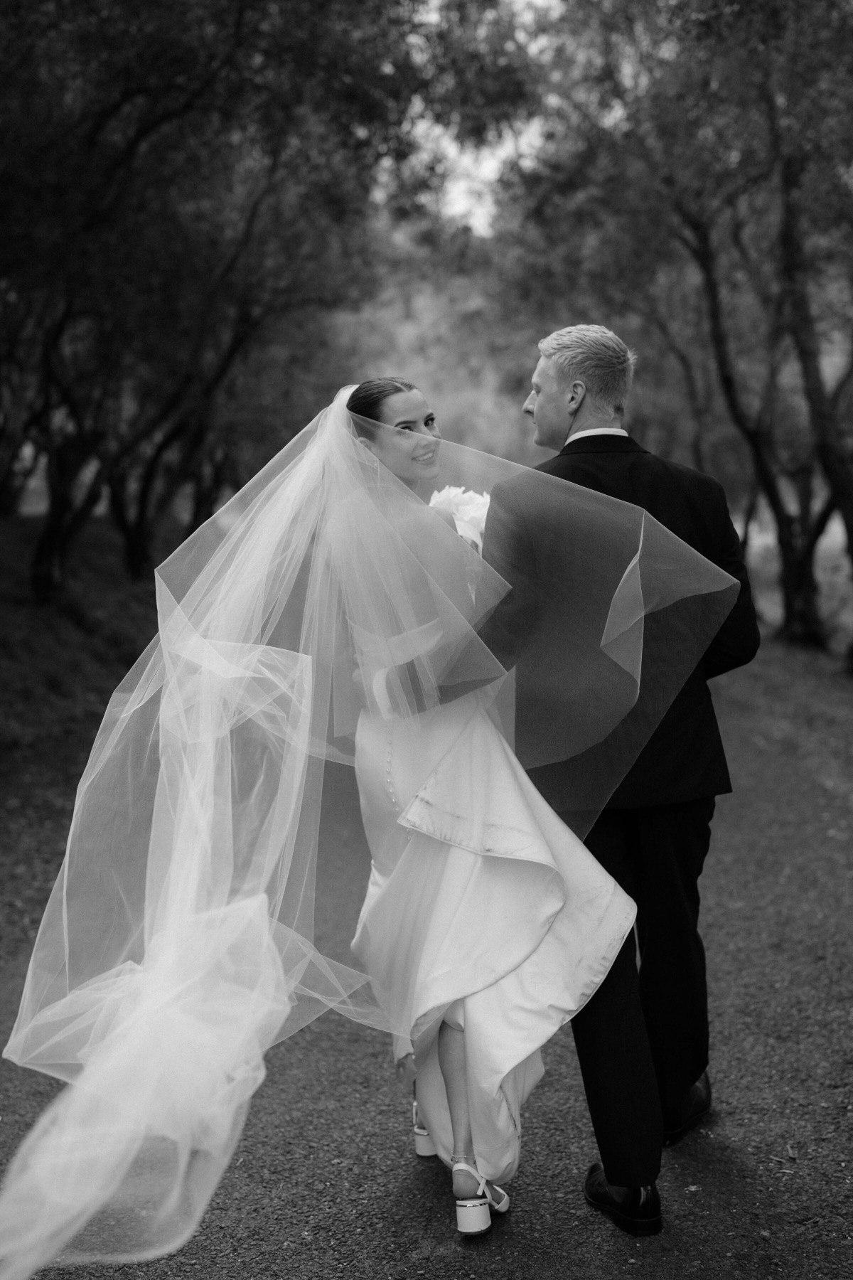 Bride's veil swirling as she walks with her groom through an olive grove